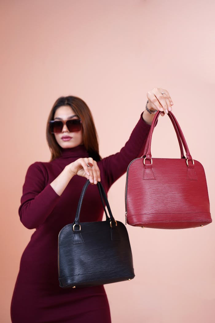 A stylish woman in sunglasses displays elegant red and black handbags in a studio shoot.
